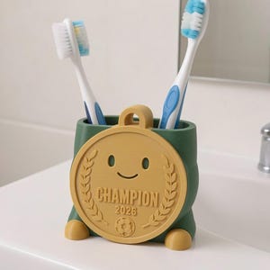 May include: A green toothbrush holder with a gold-colored medal design. The medal has a smiling face, laurel leaves, and the word "CHAMPION" with the year "2026". Two toothbrushes with blue and white bristles are inside the holder.