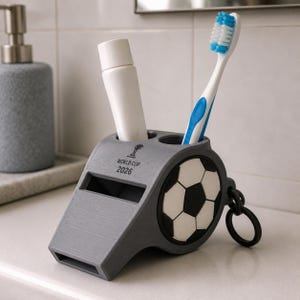 May include: A gray whistle-shaped toothbrush holder featuring a black and white soccer ball graphic on the side and the text "WORLD CUP 2026" printed on the top. The holder contains a white tube and a blue and white toothbrush.