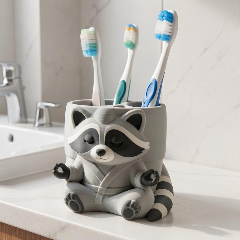May include: A grey and white raccoon-shaped toothbrush holder featuring a raccoon in a seated meditation pose with its hands in a mudra position. The holder has three circular openings at the top, currently containing three toothbrushes.