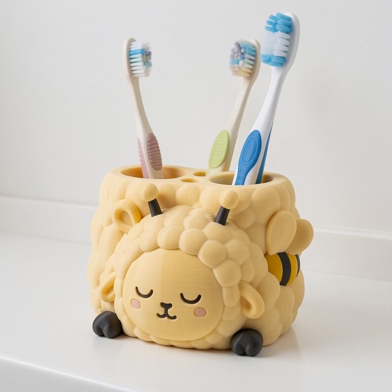 May include: A pale yellow toothbrush holder shaped like a sheep with a bee design on the side. The holder features a textured, rounded body, two black antennae, and a sleeping face with pink cheeks. Three toothbrushes are inserted into the top.