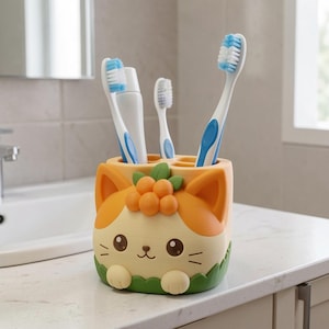 May include: A cream and orange cat-shaped toothbrush holder with green leaf accents and orange berries on top. The holder contains two blue and white toothbrushes and a white tube of toothpaste, featuring a small cat face with whiskers.