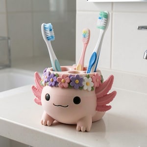 May include: A light pink axolotl-shaped toothbrush holder featuring a crown of colorful purple, pink, and white flowers around the top rim. The holder has three circular openings containing three toothbrushes with blue, pink, and green bristles.