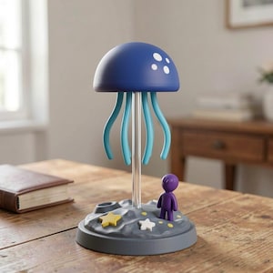 May include: A whimsical lamp featuring a blue jellyfish-shaped shade with white dots and turquoise tentacles. The lamp includes a small purple figure and star-shaped accents on a gray base, creating a playful, decorative lighting fixture.