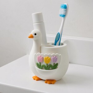 Pu&ograve; includere: Un portaspazzolino bianco a forma di oca con becco e zampe arancioni. Il corpo presenta una tasca decorativa con tre tulipani color pastello rosa, gialli e viola con steli verdi. All'interno sono inseriti un tubetto bianco e uno spazzolino blu e bianco.