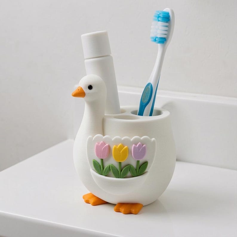 Pu&ograve; includere: Un portaspazzolino bianco a forma di oca con becco e zampe arancioni. Il corpo presenta una tasca decorativa con tre tulipani color pastello rosa, gialli e viola con steli verdi. All'interno sono inseriti un tubetto bianco e uno spazzolino blu e bianco.