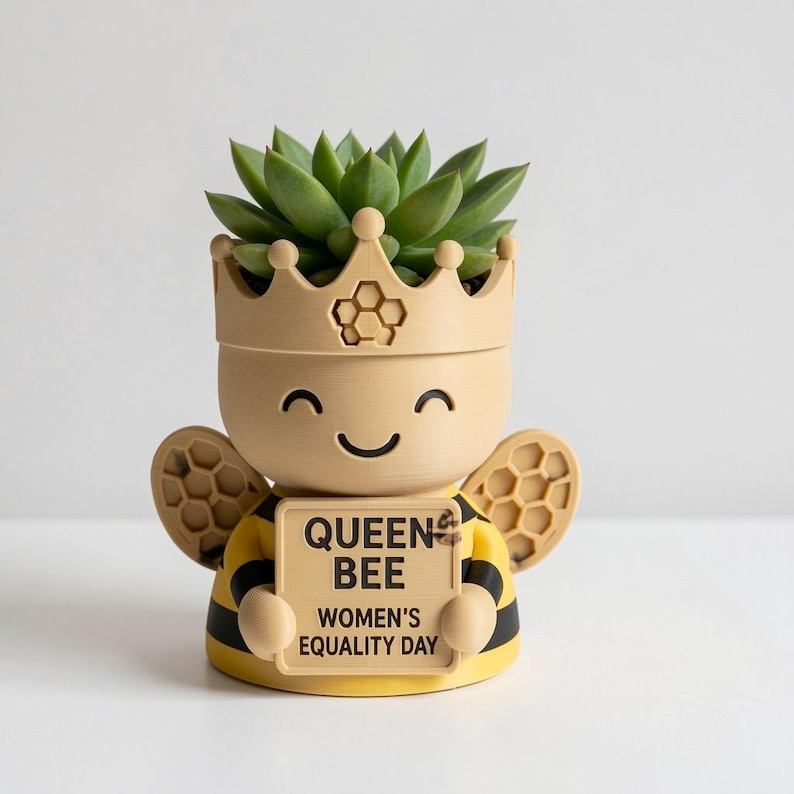 May include: A beige and yellow bee-shaped planter with a crown top, holding a sign that reads QUEEN BEE WOMEN'S EQUALITY DAY. The planter features black stripes, honeycomb-patterned wings, and a smiling face, containing a green succulent plant.