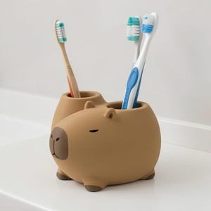 May include: A tan capybara-shaped toothbrush holder with a dark brown nose and feet. The holder features two circular openings on the top, currently containing one tan toothbrush and two white toothbrushes with blue and green bristles.