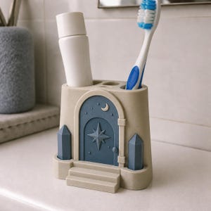 Pu&ograve; includere: Un portaspazzolino beige a forma di edificio con una porta ad arco blu, una stella e una mezzaluna. Il supporto presenta due pilastri blu verticali e tre gradini alla base. Contiene un tubetto bianco e uno spazzolino da denti blu e bianco.