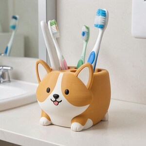 Puede incluir: Un soporte para cepillos de dientes con forma de Corgi en color tostado y blanco, con una nariz negra y una peque&ntilde;a lengua. El soporte tiene cuatro aberturas circulares en la parte superior que contienen cuatro cepillos de dientes con cerdas y mangos azules, verdes y blancos.