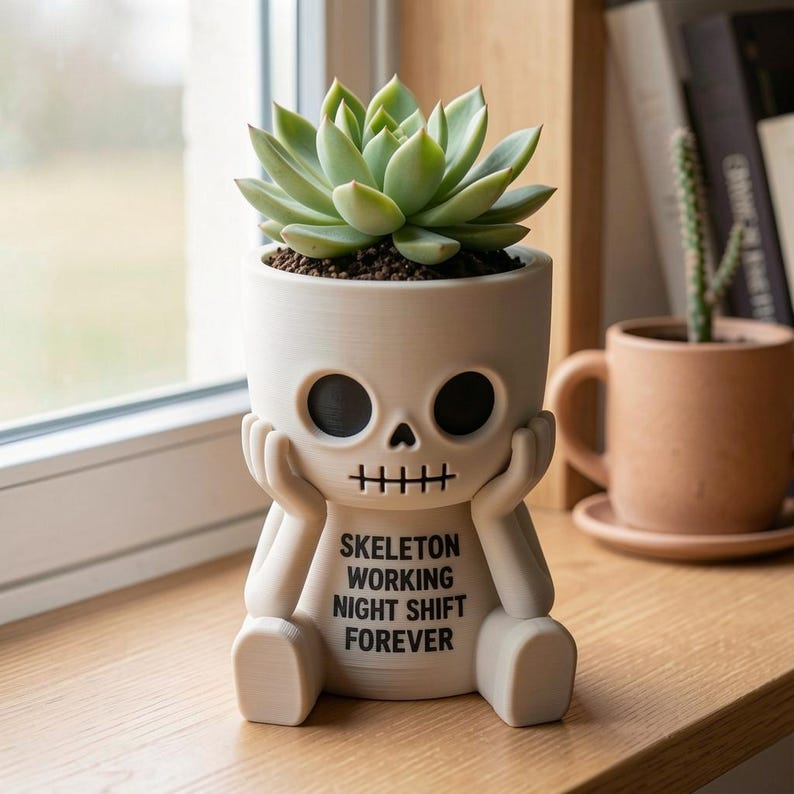 May include: A matte off-white planter shaped like a skeleton resting its chin on its hands. The front features black text that reads SKELETON WORKING NIGHT SHIFT FOREVER. A green succulent plant is potted inside the hollow head of the skeleton figure.