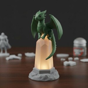 May include: A green dragon figurine with outstretched wings sits atop a glowing, faceted crystal. The crystal is mounted on a gray, rocky base. The dragon has a long tail and is a fantasy-themed decorative item.