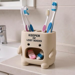 May include: A beige, character-shaped toothbrush holder with the text KEEPER OF LOST SOCKS printed on the front. The holder features a hollow mouth area containing two small, colorful socks and holds three toothbrushes with blue and pink bristles.