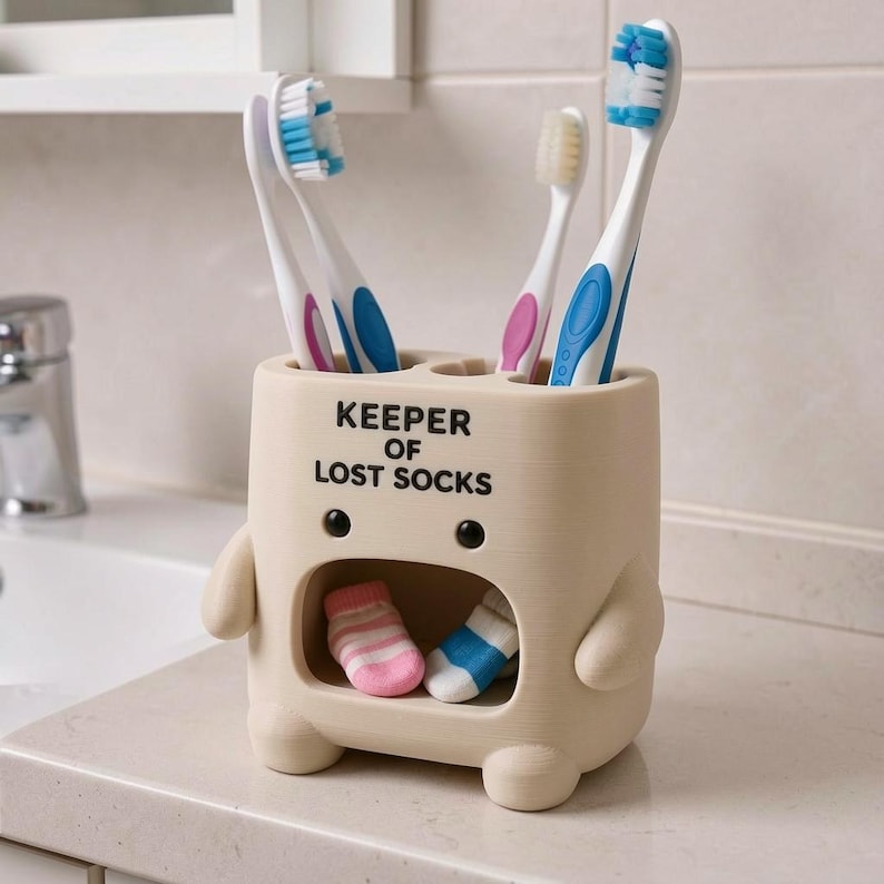 May include: A beige, character-shaped toothbrush holder with the text KEEPER OF LOST SOCKS printed on the front. The holder features a hollow mouth area containing two small, colorful socks and holds three toothbrushes with blue and pink bristles.