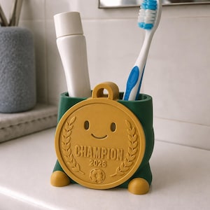 May include: A green toothbrush holder with a gold medal design. The medal has a smiling face, laurel wreath, and the word "CHAMPION" with the year "2026". A tube of toothpaste and a toothbrush are inside the holder.