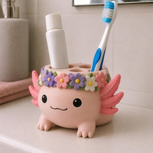 May include: A light pink axolotl-shaped toothbrush holder featuring a crown of purple, pink, and white flowers. The holder has four circular openings and contains a white tube of toothpaste and a blue and white toothbrush with soft bristles.
