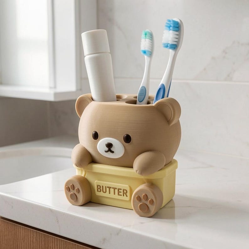 Puede incluir: Un soporte para cepillos de dientes con forma de oso de color marr&oacute;n claro y una base rectangular amarilla con la palabra BUTTER grabada en el frente. El soporte contiene un tubo blanco y dos cepillos de dientes con cerdas azules y blancas.