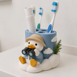 Puede incluir: Un soporte cil&iacute;ndrico azul para cepillos de dientes con un pato de dibujos animados que lleva un sombrero beige y una chaqueta azul, sosteniendo binoculares negros. Tiene nubes blancas, una ramita verde, un tubo de pasta dental y tres cepillos.