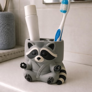 May include: A grey raccoon-shaped toothbrush holder featuring a raccoon in a seated meditation pose with its eyes closed. The holder has two cylindrical openings at the top, currently containing a white tube and a blue and white toothbrush.