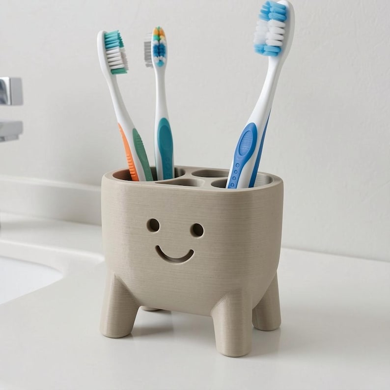 Pu&ograve; includere: Un portaspazzolini beige con un design a faccina sorridente, che contiene tre spazzolini con accenti blu, verdi e arancioni. Il supporto ha quattro scomparti e quattro piccole gambe ed &egrave; appoggiato su una superficie bianca.