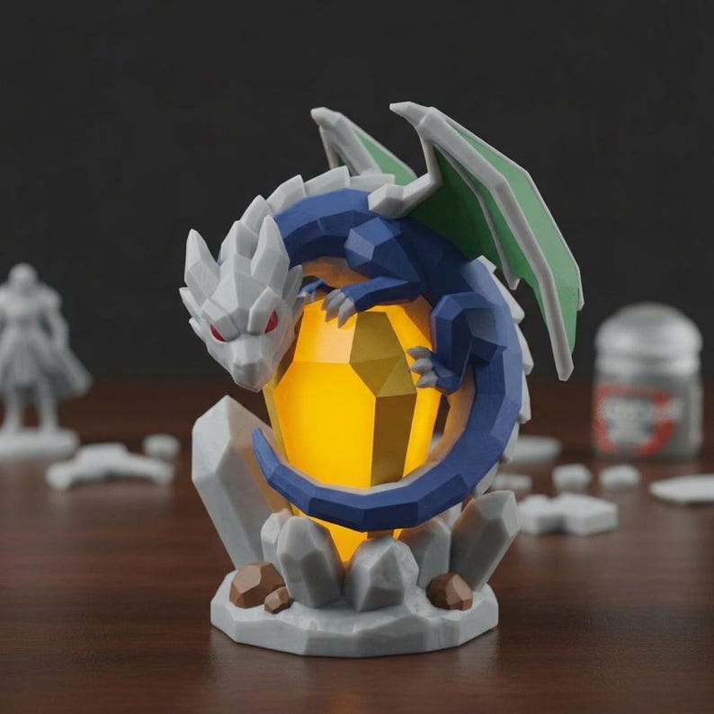 Puede incluir: Una figura geom&eacute;trica de drag&oacute;n en tonos azules, blancos y verdes, posada sobre un cristal amarillo brillante. El drag&oacute;n tiene ojos rojos y est&aacute; rodeado de formaciones rocosas grises. Un art&iacute;culo decorativo de tem&aacute;tica fant&aacute;stica.