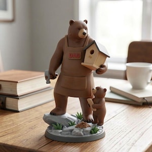 K&ouml;nnte beinhalten: Eine braune B&auml;renfigur mit einer braunen Sch&uuml;rze mit dem Wort "DAD" darauf, die ein Vogelhaus und einen Hammer h&auml;lt. Eine kleinere B&auml;renfigur steht neben dem gr&ouml;&szlig;eren B&auml;ren und h&auml;lt ein St&uuml;ck Holz. Die Figuren stehen auf einem Sockel mit Felsen und Gras.