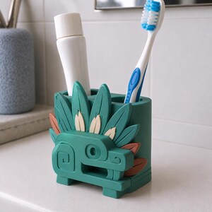 May include: A dark green toothbrush holder featuring a stylized serpent design with raised leaf-like details in green, tan, and brown. The holder has two cylindrical compartments, one containing a white tube and the other a blue and white toothbrush.
