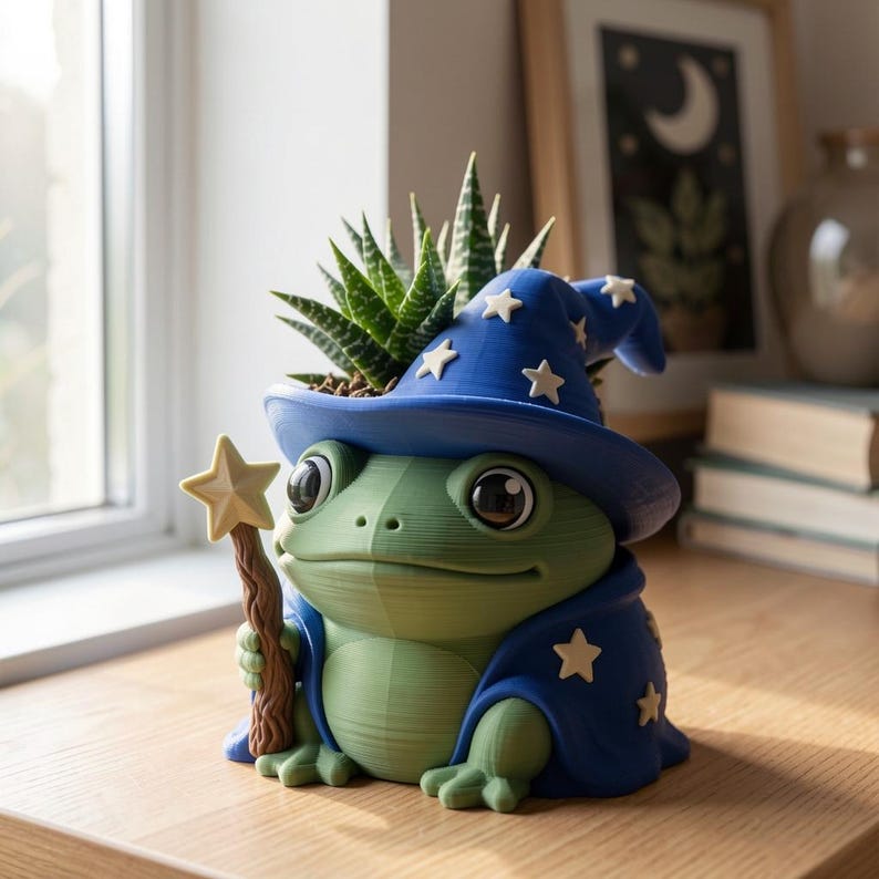 May include: A green frog planter wearing a blue wizard hat and cape decorated with white stars. The frog holds a brown wooden-textured wand topped with a white star. A green succulent plant grows from the top of the hat, which serves as the planter.