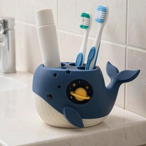 May include: A dark blue and white whale-shaped toothbrush holder featuring a small yellow planet graphic on the side, star-shaped cutouts, and three circular openings holding a white tube and two toothbrushes with blue and green bristles.