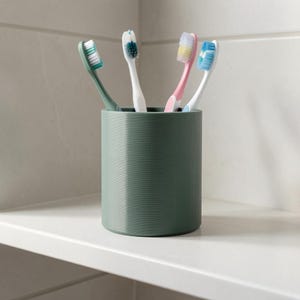 Pu&ograve; includere: Un portaspazzolini cilindrico color verde salvia contenente quattro spazzolini. Gli spazzolini hanno manici bianchi e setole di vari colori, tra cui verde, blu scuro, rosa e azzurro, tutti posizionati verticalmente nel contenitore.