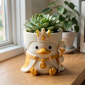May include: A white and yellow planter shaped like a duck wearing a crown and cape, holding a small scepter. The duck has a round pendant featuring a smaller duck face. A green succulent plant is potted inside the top of the duck-shaped container.