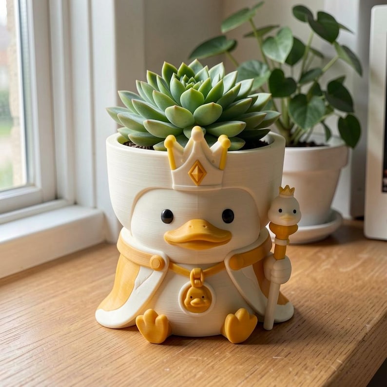 May include: A white and yellow planter shaped like a duck wearing a crown and cape, holding a small scepter. The duck has a round pendant featuring a smaller duck face. A green succulent plant is potted inside the top of the duck-shaped container.