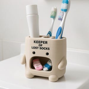 May include: A beige, creature-shaped toothbrush holder with the text KEEPER OF LOST SOCKS printed in black. The holder features two small arms, two black eyes, and a wide mouth containing two miniature socks. It holds a white tube and two toothbrushes.