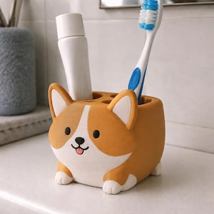 Puede incluir: Un soporte para cepillos de dientes con forma de perro en color canela y blanco, con cara de dibujos animados, nariz negra, lengua rosa y dos orejas erguidas. El soporte contiene un tubo de pasta dental blanco y un cepillo azul y blanco.
