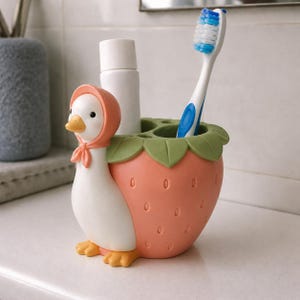 May include: A white and orange duck-shaped toothbrush holder with a pink bonnet, attached to a pink strawberry-shaped base with green leaves at the top. The holder contains a white tube and a blue and white toothbrush.