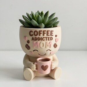 May include: A cream-colored planter shaped like a seated figure holding a small pink mug with a heart design. The planter features the raised brown and pink text COFFEE ADDICTED MOM, accented with small pink hearts and brown coffee bean shapes.
