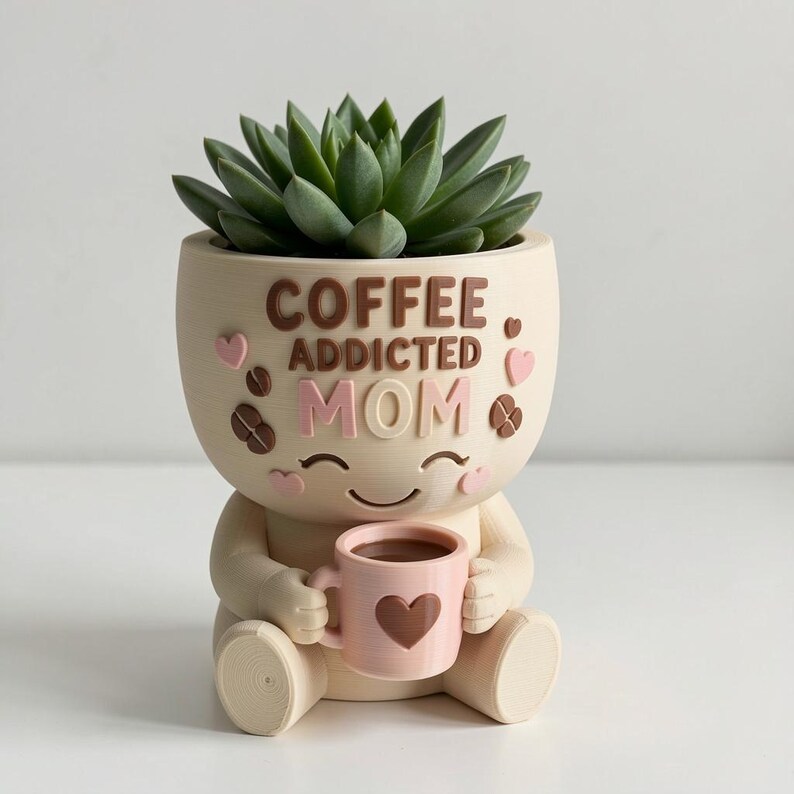 May include: A cream-colored planter shaped like a seated figure holding a small pink mug with a heart design. The planter features the raised brown and pink text COFFEE ADDICTED MOM, accented with small pink hearts and brown coffee bean shapes.