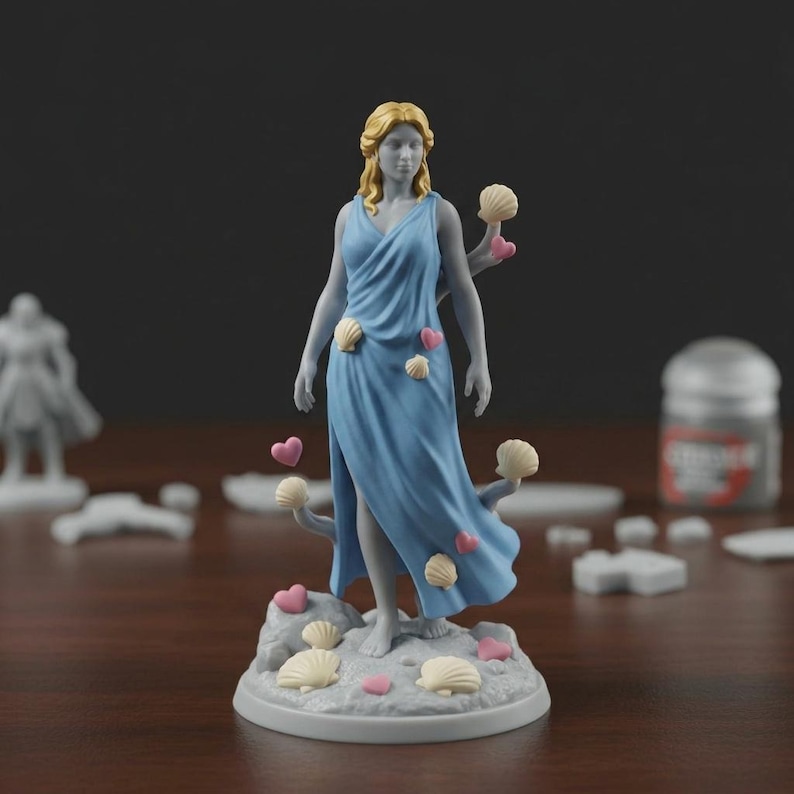 May include: A miniature figurine of a woman in a flowing blue dress, adorned with seashell and heart accents. The figure has blonde hair and stands on a base with more seashells and hearts. The figurine is likely for tabletop gaming or display.