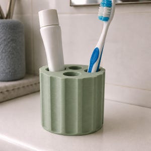 May include: A sage green toothbrush holder with a tube of toothpaste and a toothbrush. The holder has a cylindrical shape with vertical grooves and three circular openings. The toothbrush is white with blue accents. The toothpaste tube is white.