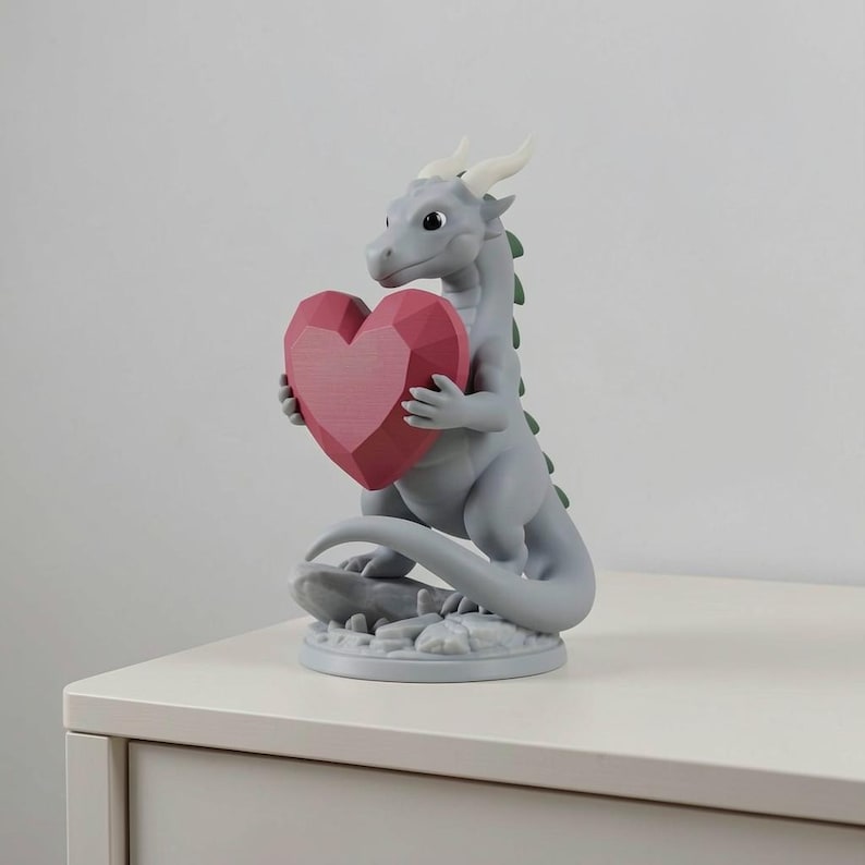 Peut inclure: Figurine de dragon gris tenant un c&oelig;ur g&eacute;om&eacute;trique rouge. Le dragon a des cornes blanches et des &eacute;pines vertes, pos&eacute; sur une base ronde. Objet d&eacute;coratif, possiblement pour la Saint-Valentin ou une occasion similaire.