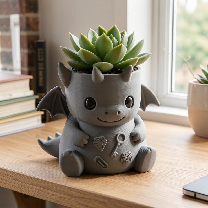 May include: A matte gray dragon-shaped planter featuring a succulent plant. The dragon has small wings, horns, and a tail, with embossed icons of a diamond, a key, a paperclip, and a twenty-sided die on its front. The planter has a smooth finish.