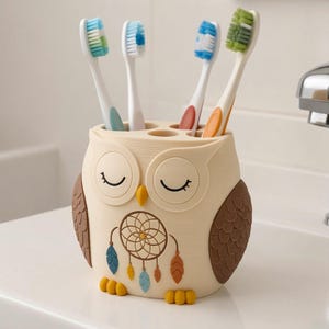 May include: A cream-colored owl-shaped toothbrush holder with brown wings and yellow feet. The owl features closed eyes, a yellow beak, and a dreamcatcher graphic with blue, yellow, and orange feathers. Four toothbrushes are inserted inside.