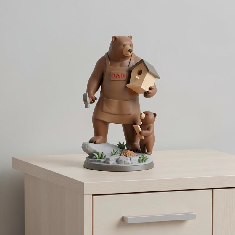 K&ouml;nnte beinhalten: Eine braune B&auml;renfigur mit Hammer und Vogelhaus. Der B&auml;r tr&auml;gt eine braune Sch&uuml;rze mit dem Wort "DAD" und steht mit einem kleineren B&auml;ren, der ein Holzbrett h&auml;lt. Die Figur steht auf einem grauen Sockel mit gr&uuml;nem Gras und kleinen Steinen.