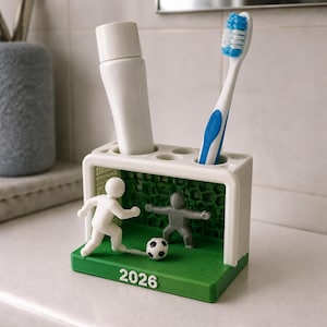 May include: A white and green plastic toothbrush holder shaped like a football goal with a white player figure, a grey goalkeeper figure, a small football, and the text 2026 on the front. It features multiple circular slots for toiletries.