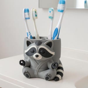 May include: A grey, cylindrical toothbrush holder shaped like a raccoon sitting in a meditative pose with its hands in a mudra position. The raccoon has a black mask, a striped tail, and is wearing a robe. Four toothbrushes are stored inside.