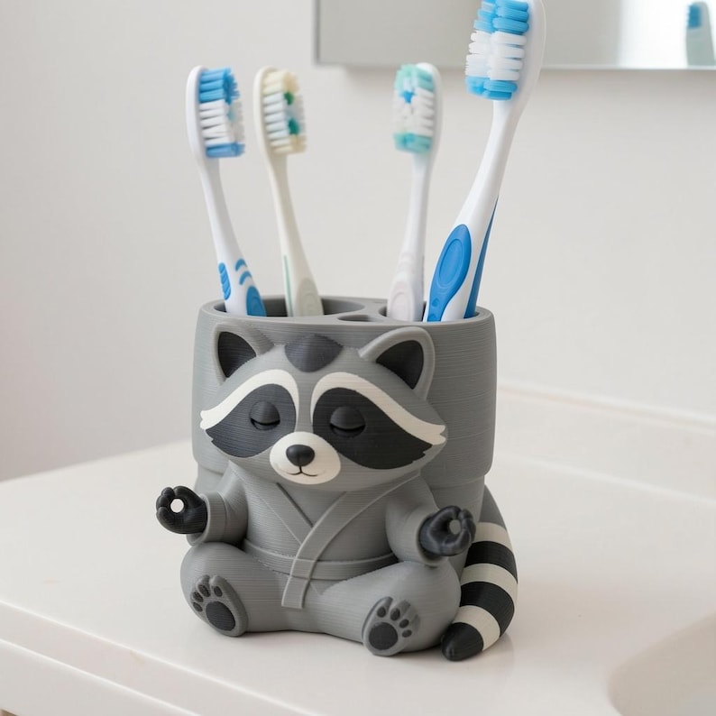 May include: A grey, cylindrical toothbrush holder shaped like a raccoon sitting in a meditative pose with its hands in a mudra position. The raccoon has a black mask, a striped tail, and is wearing a robe. Four toothbrushes are stored inside.
