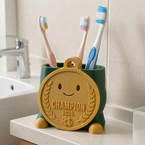 May include: A green toothbrush holder with a gold medal design. The medal has a smiling face, laurel leaves, and the word "CHAMPION" with the year "2026". Three toothbrushes with different colored bristles are inside.