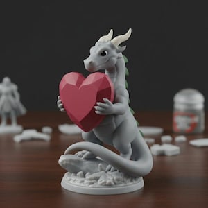 Peut inclure: Figurine de dragon gris tenant un c&oelig;ur rouge facett&eacute;. Le dragon a des cornes blanches, des accents verts et est perch&eacute; sur une base rocheuse. La figurine est sur une base ronde.