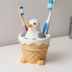 May include: A toothbrush holder shaped like a white and gray seagull sitting in a tan waffle cone base. The holder features two small feet at the bottom and holds two toothbrushes, one with a blue and white handle and one with a tan handle.