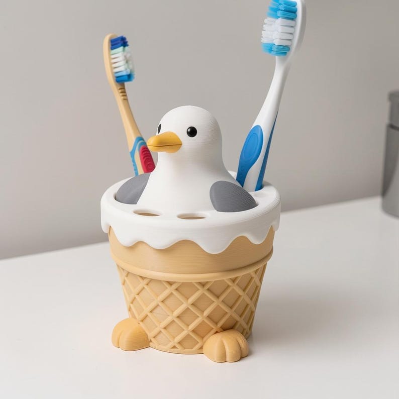 May include: A toothbrush holder shaped like a white and gray seagull sitting in a tan waffle cone base. The holder features two small feet at the bottom and holds two toothbrushes, one with a blue and white handle and one with a tan handle.