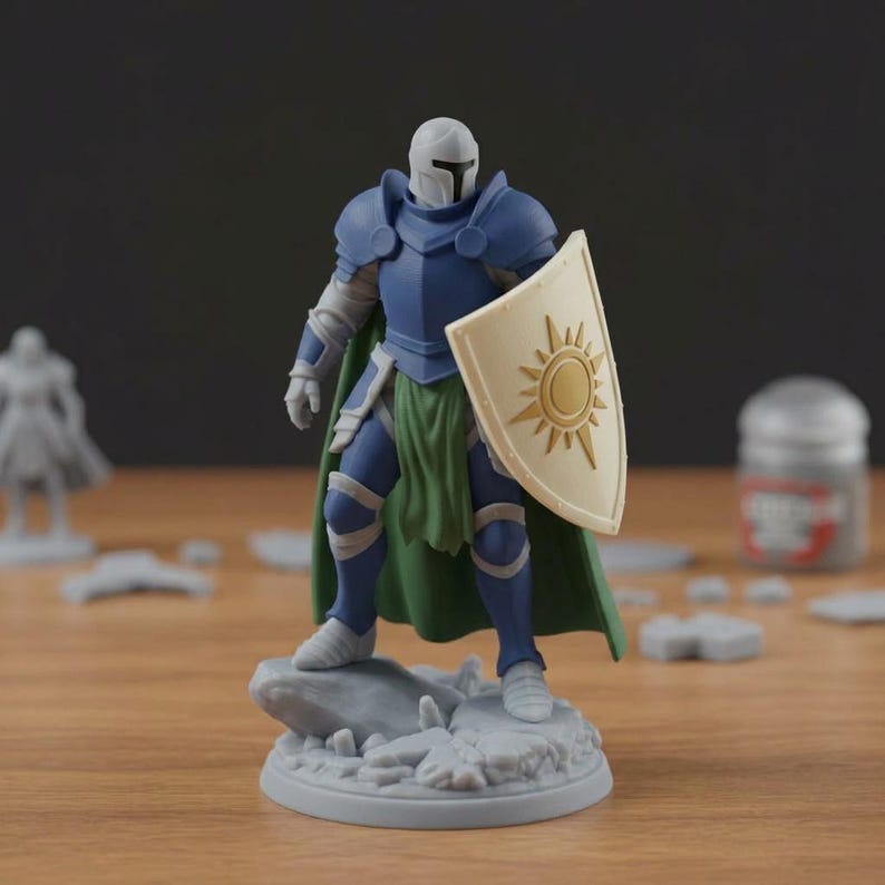 May include: A miniature figurine of a knight in blue armor, a white helmet, and a green cape. The knight holds a shield with a sun design. The figurine is on a gray rocky base, with other miniature pieces in the background.